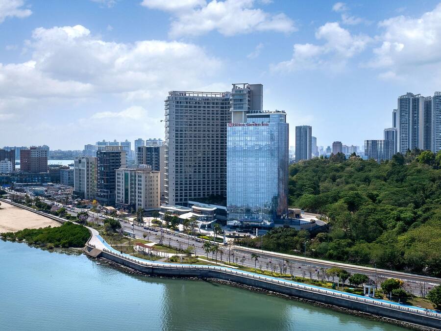 Un hotel moderno de altura con fachada de vidrio, ubicado cerca de una orilla con un puente y vegetación.