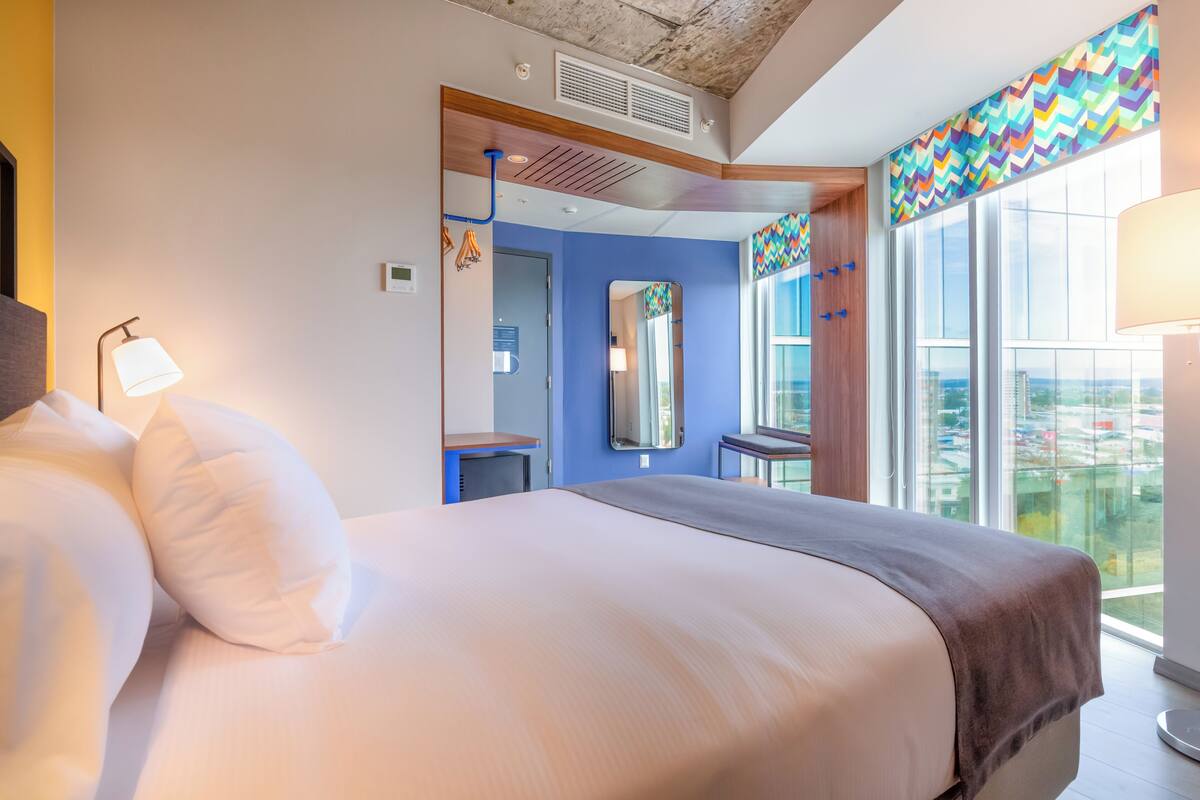 A modern hotel room with a large bed, a desk, and a view of the city through large windows.