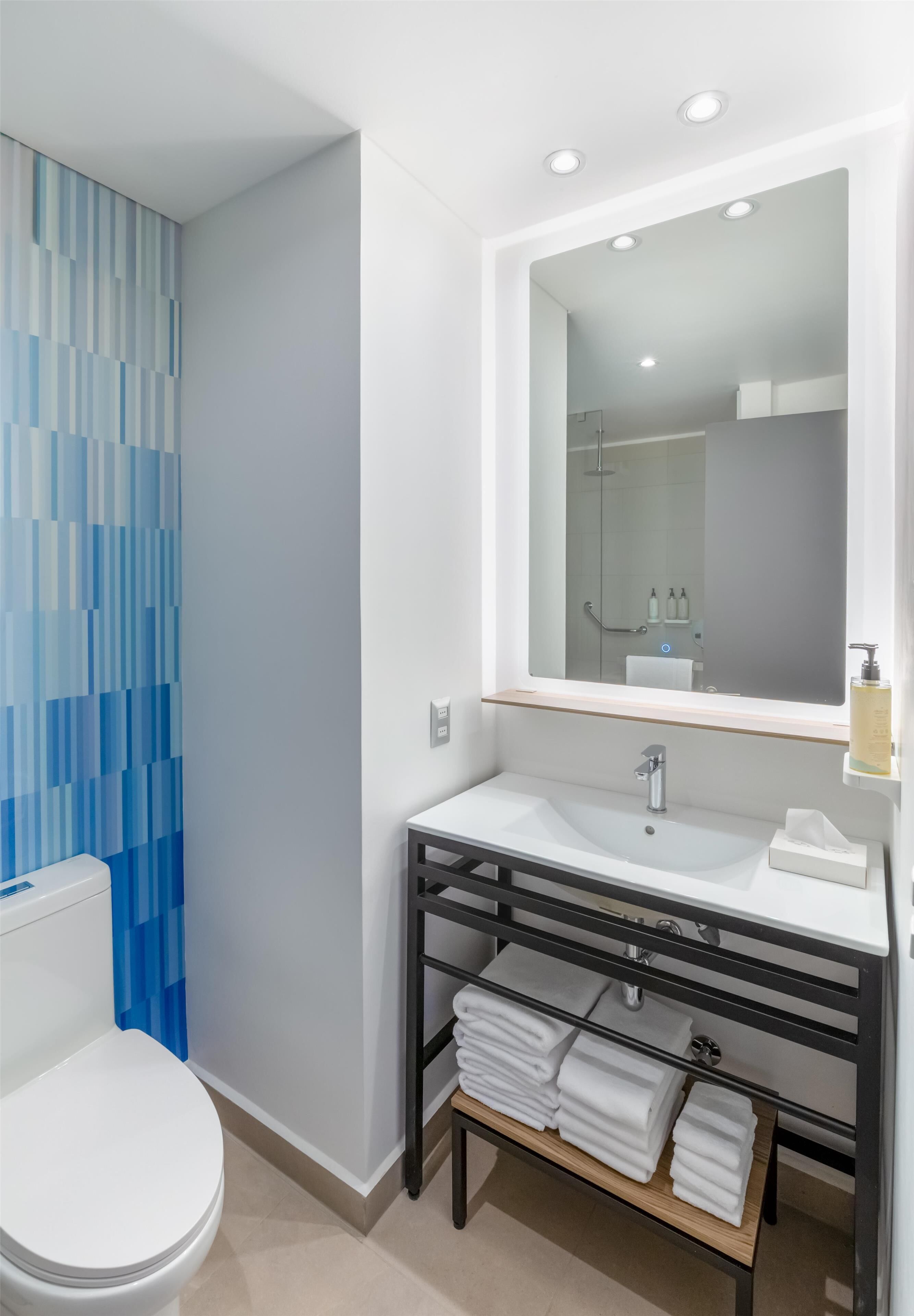 Un baño moderno con un lavamanos blanco, un espejo grande y un inodoro. Una pared con diseño azul y un estante con toallas.