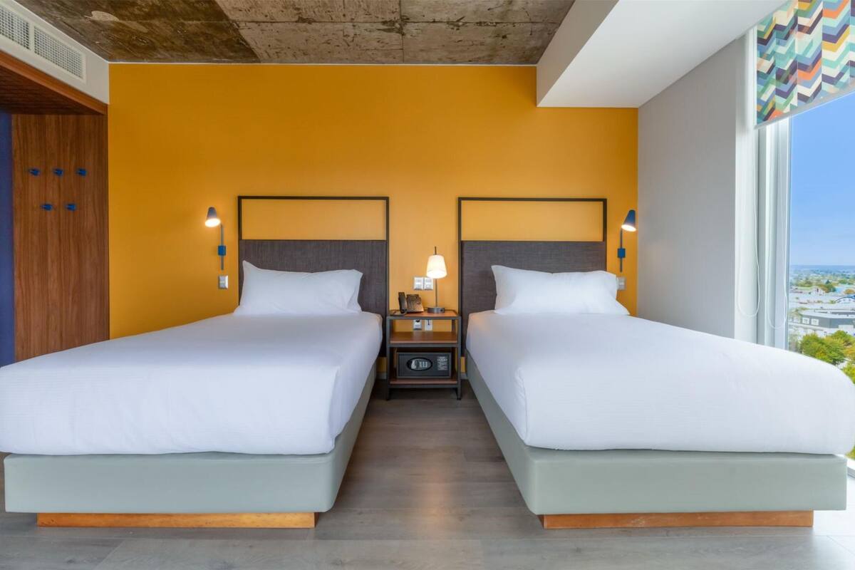 A hotel room with two beds, a nightstand with a lamp, and a view of the city.