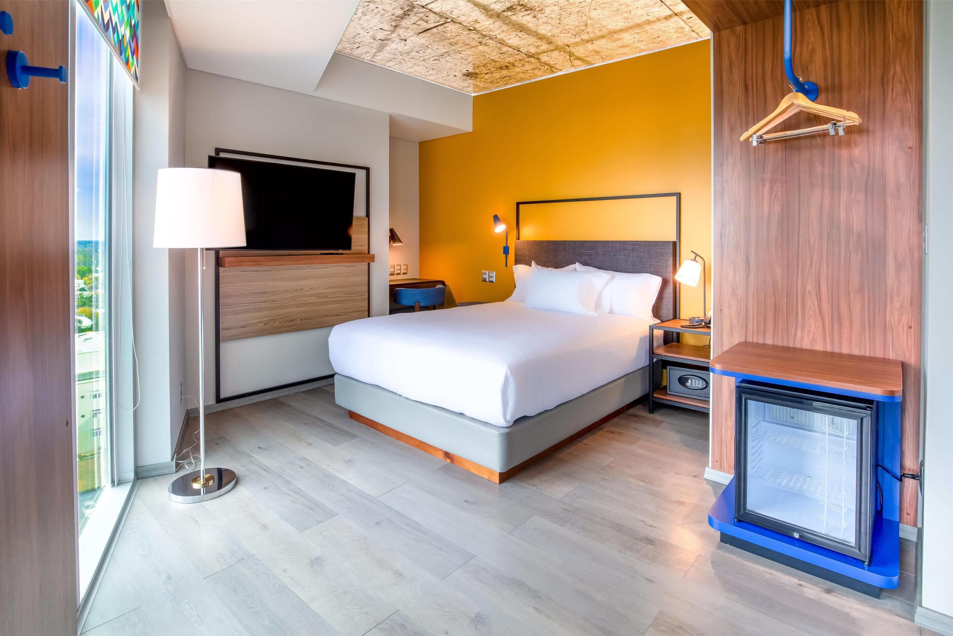 Moderni hotellihuone, jossa on sänky, yöpöytä, lamppu ja sisäänrakennettu minijääkaappi.