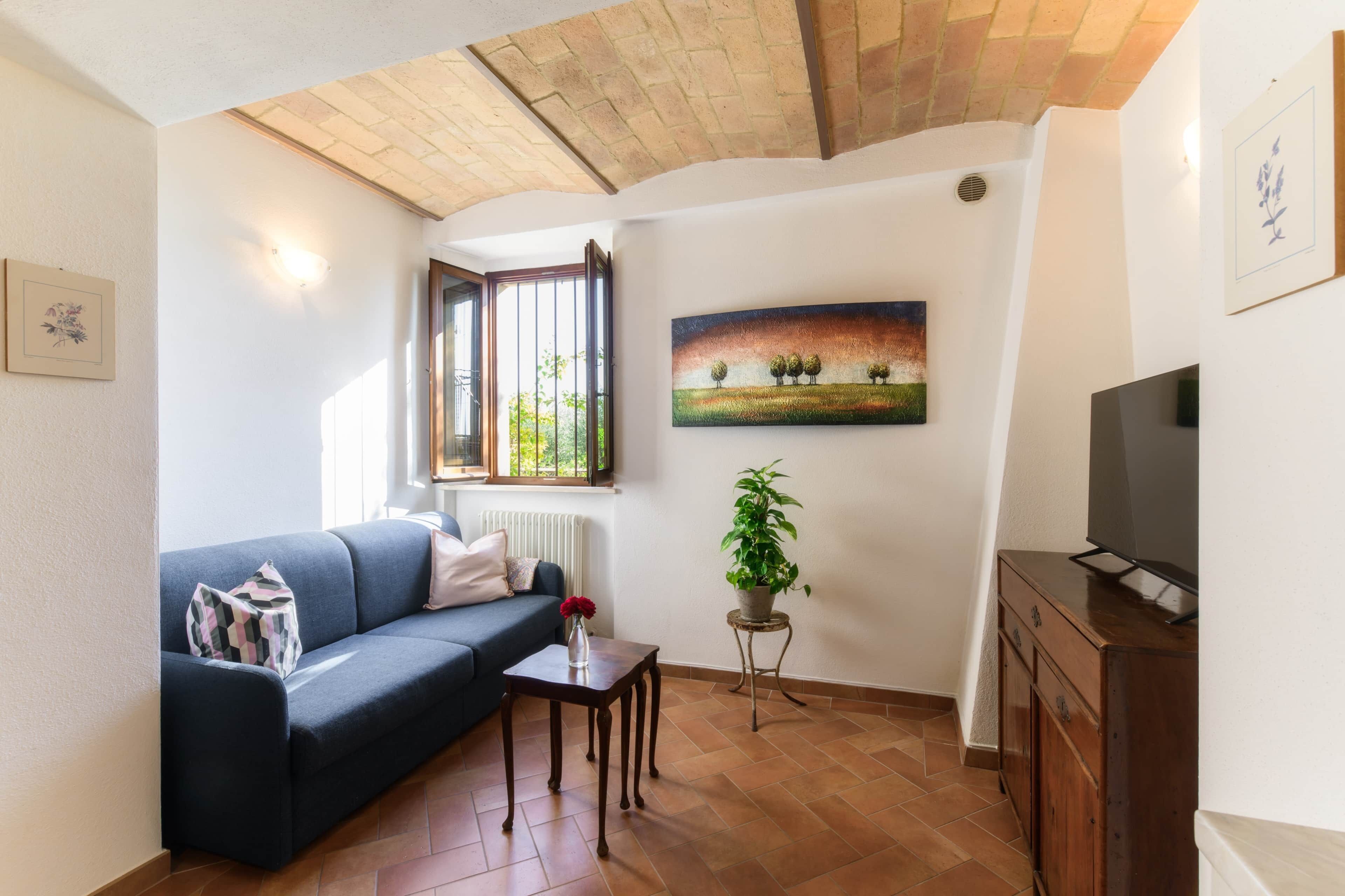 Un salon avec un canapé, une petite table sur laquelle est posé un vase, une plante en pot, un tableau et un téléviseur.
