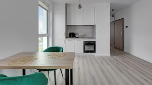 A modern kitchen with white cabinets, a built-in oven, and a dining area with a wooden table and green chairs. - Studio Sw. Piotra by Grand Apartments (Gdynia)