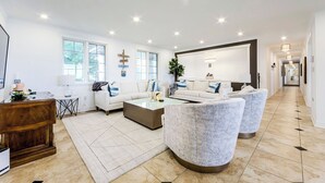 Un salon spacieux avec un canapé blanc, une table basse et un grand téléviseur. De nombreuses fenêtres laissent entrer la lumière naturelle et offrent une vue sur l’extérieur.