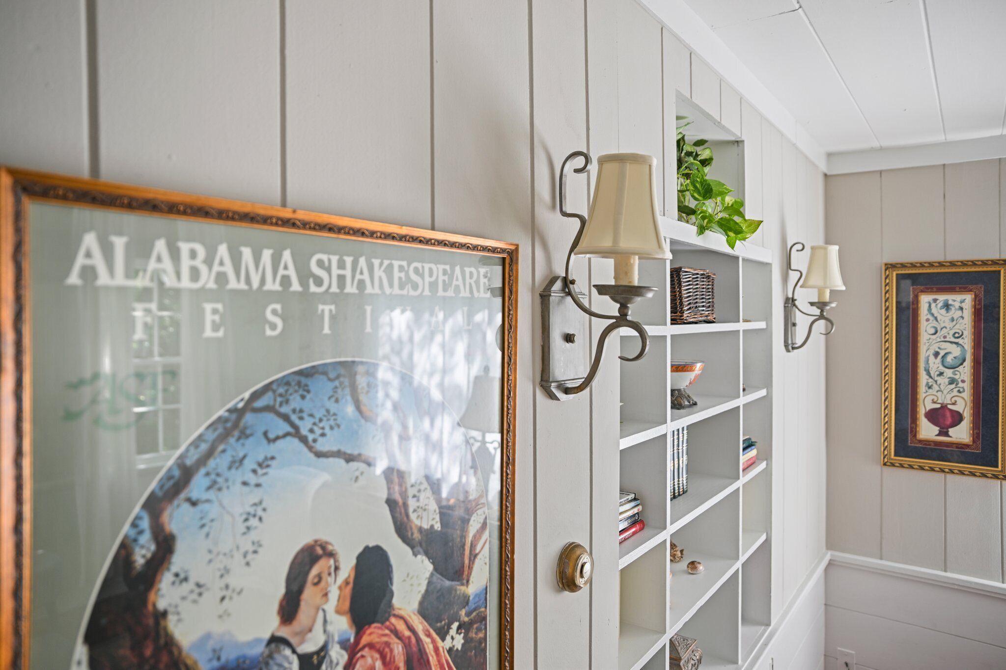 A hallway with a framed poster of 'Alabama Shakespeare Festival', a white shelving unit with decorative items, and wall-mounted lamps.