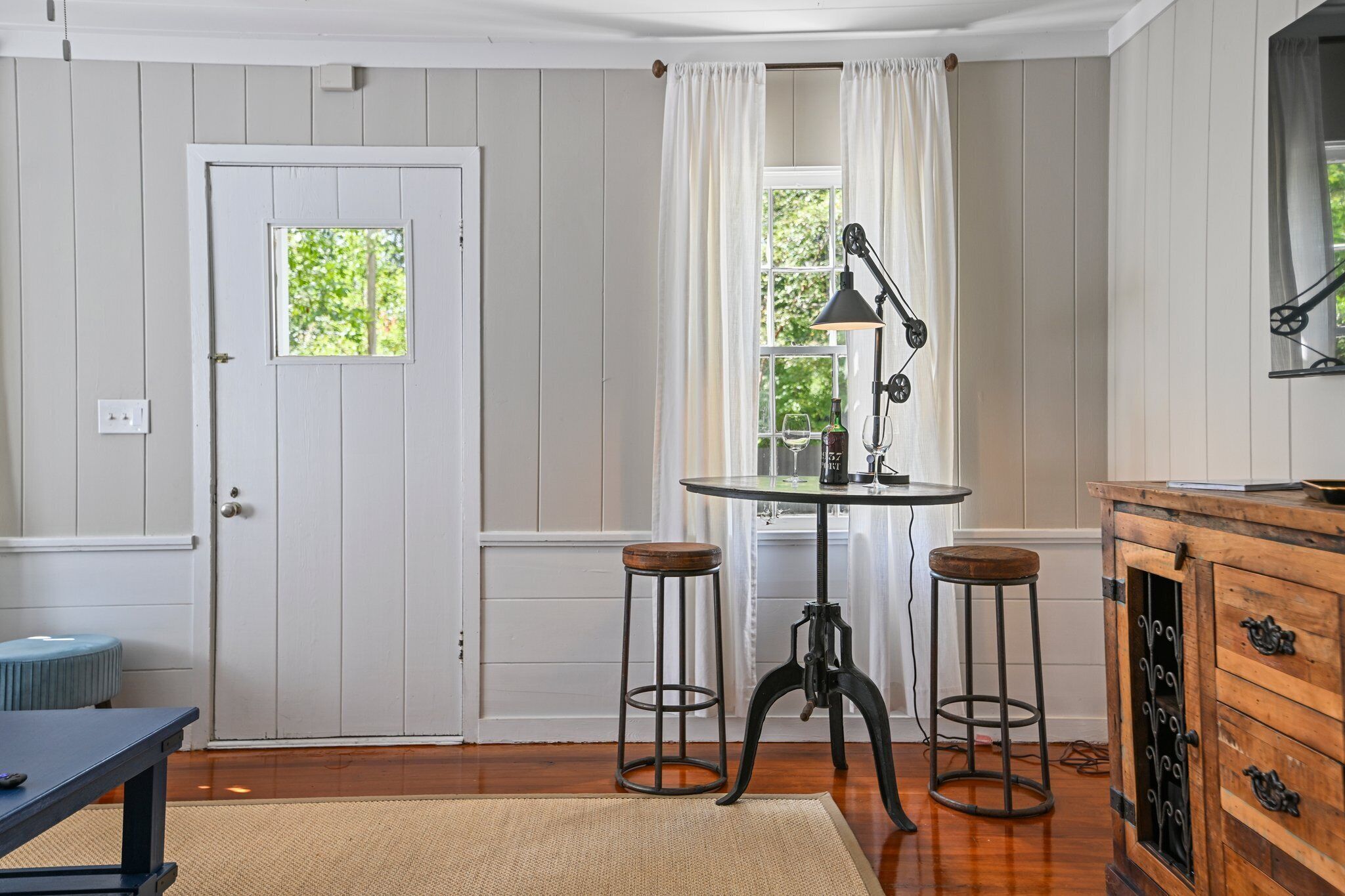 A room with a white door, a window with white curtains, a round table with two stools, a wooden cabinet with drawers, and a small stool by the door.