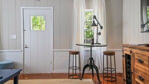 Een kamer met een witte deur, een raam met witte gordijnen, een ronde tafel met twee krukken, een houten kast met lades, en een klein zitje bij de deur.