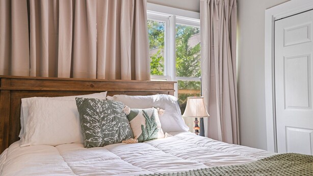 Een net opgemaakt bed met witte lakens en decoratieve kussens, een houten hoofdbord en uitzicht op groen buiten door een raam met beige gordijnen.