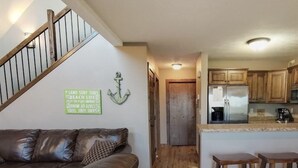 Ein geräumiges Wohnzimmer mit einer braunen Ledersofa und Holzböden. Im Hintergrund ist eine Küche mit Holzschränken und einem Edelstahlkühlschrank erkennbar.