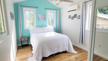 Un dormitorio con cama, dos ventanas, ventilador de techo y piso de madera.