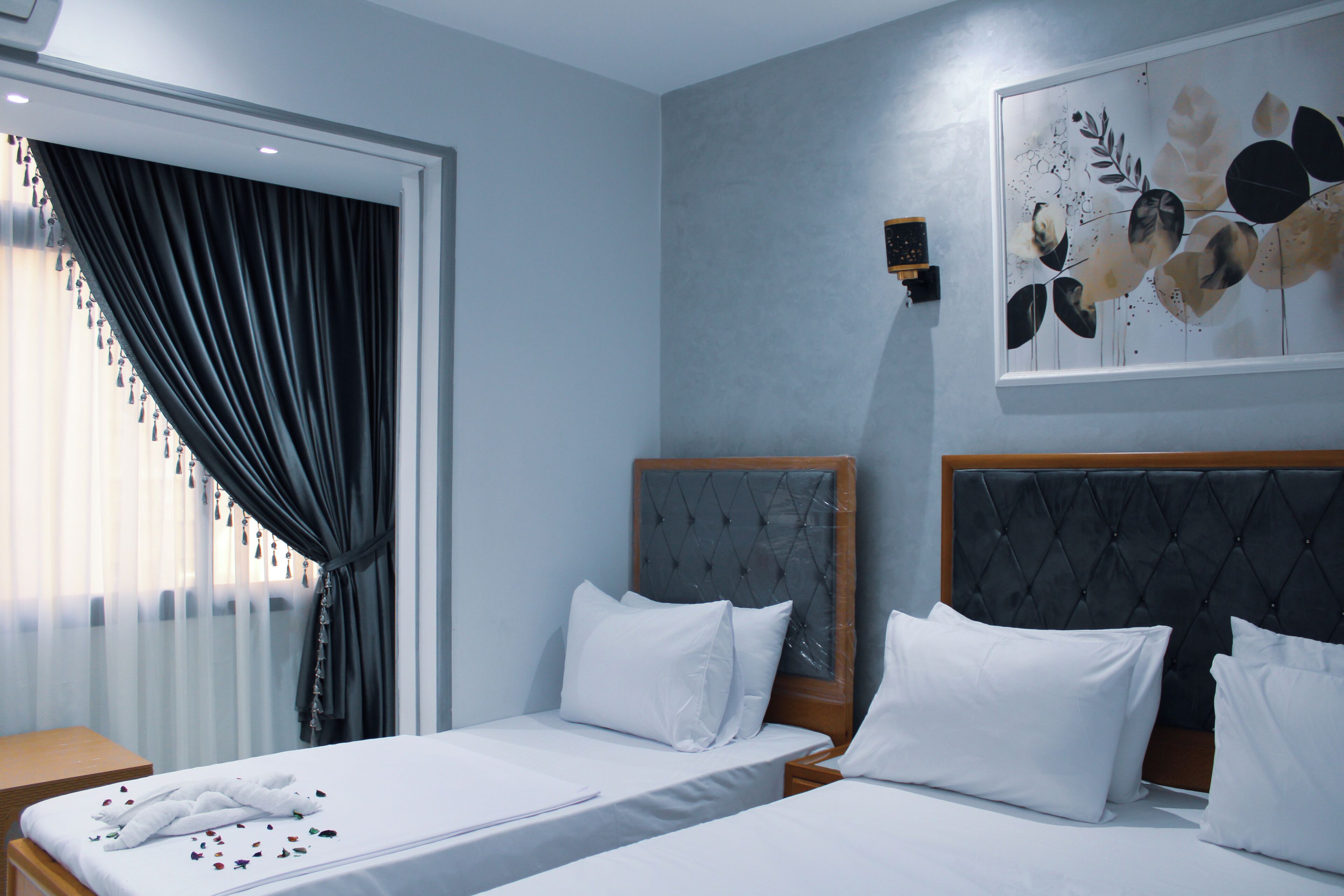 A hotel room with two beds, a large window with sheer curtains, a wall-mounted artwork, and a wooden nightstand.