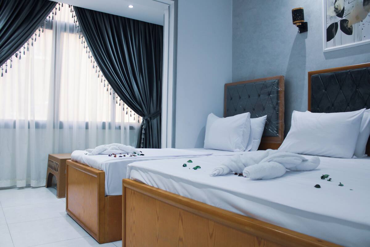 Ein Hotelzimmer mit zwei Betten, Holz-Kopfteilen, weißer Bettwäsche und einem großen Fenster mit durchsichtigen Vorhängen.