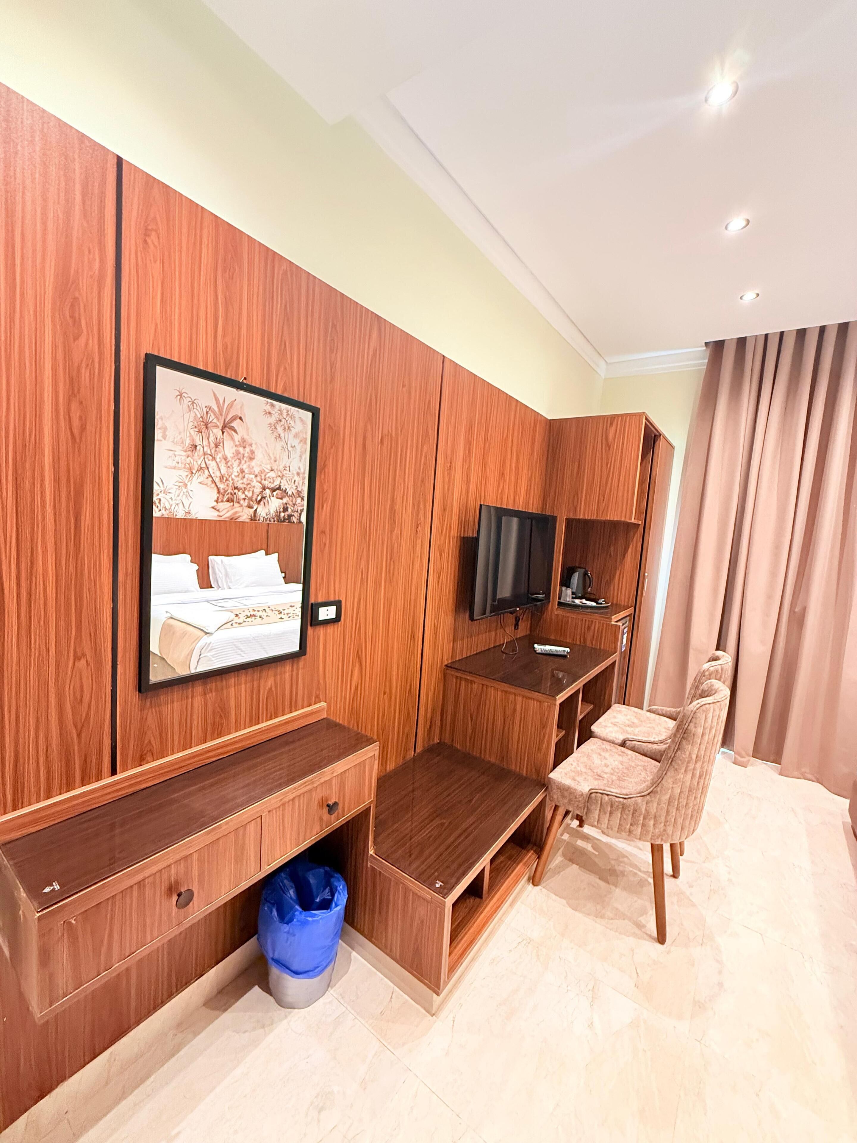 Una habitación de hotel con un armario de madera, una cama con cabecera, un televisor, un escritorio con silla y un cubo de basura azul.