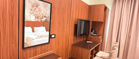 Una habitación de hotel con un armario de madera, una cama con cabecera, un televisor, un escritorio con silla y un cubo de basura azul.