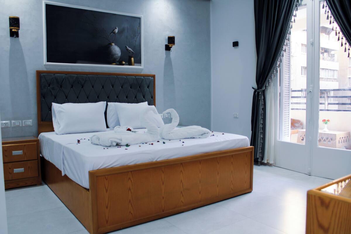 Ein Schlafzimmer mit einem großen Bett, einem Kopfteil, einem Nachttisch, einem Wandbild und einem Fenster mit Vorhängen.