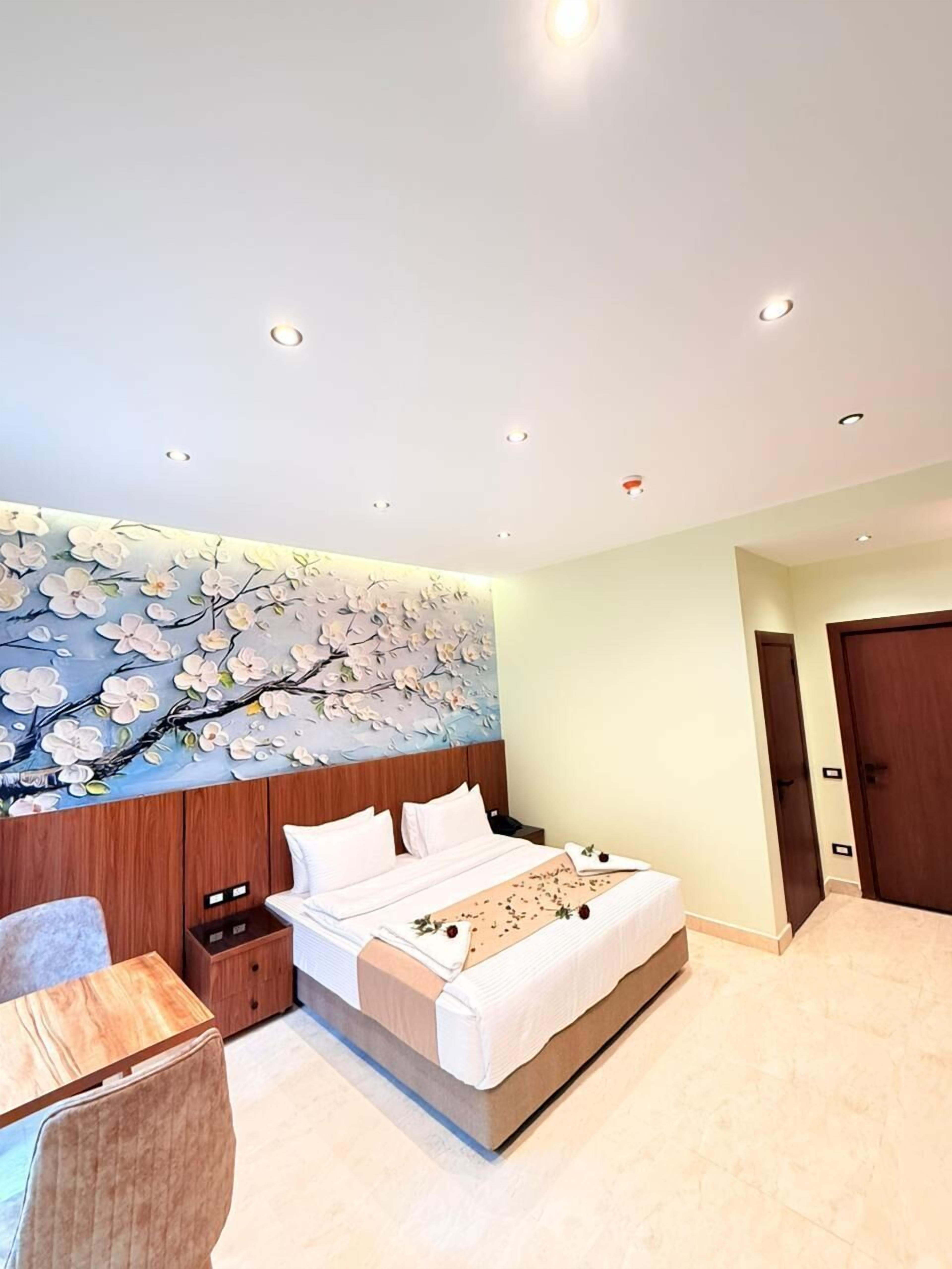 A bedroom with a large bed, a wooden nightstand, a desk, and a wall with a floral mural.