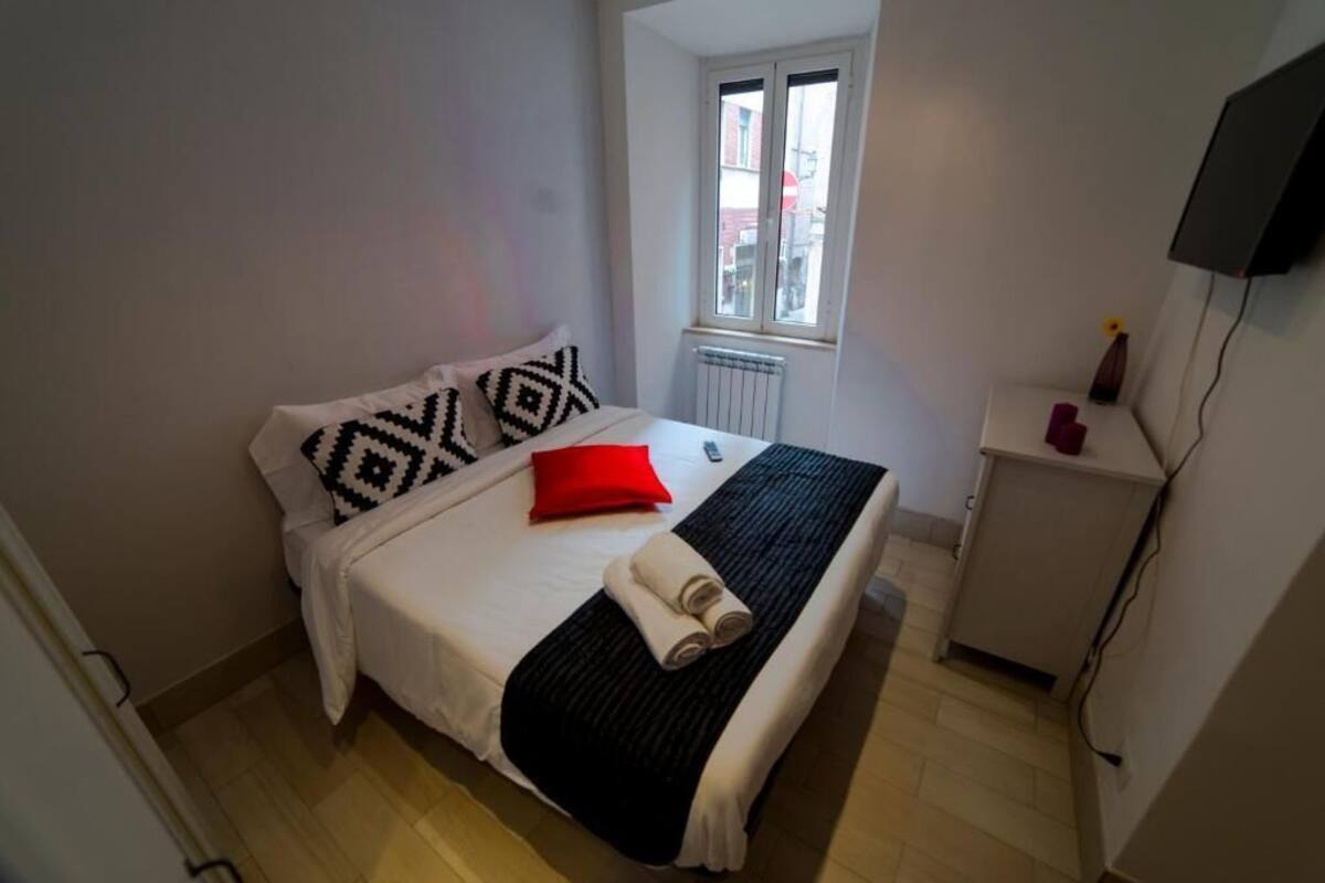 A neatly made bed with patterned pillows, a red accent pillow, and rolled towels. A small table with a vase and a candle is beside the bed.