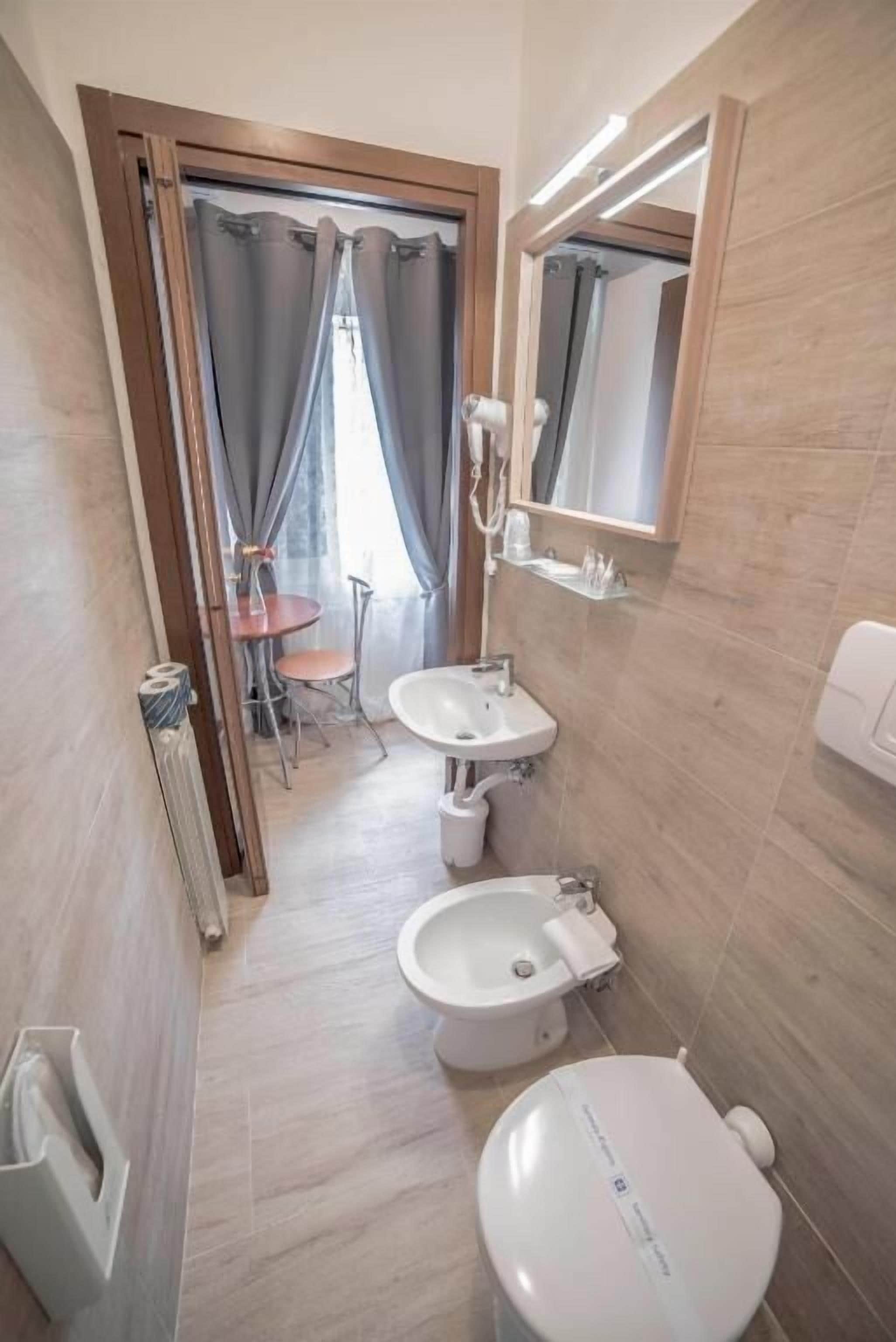 Ein Badezimmer mit zwei Waschbecken, einer Toilette und einem Spiegel. Im angrenzenden Raum befindet sich ein kleiner Tisch mit Stühlen.
