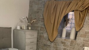 Une chambre à coucher avec une couvre-lit à motifs géométriques, une commode en bois, une fenêtre avec un rideau marron et un mur en pierre.