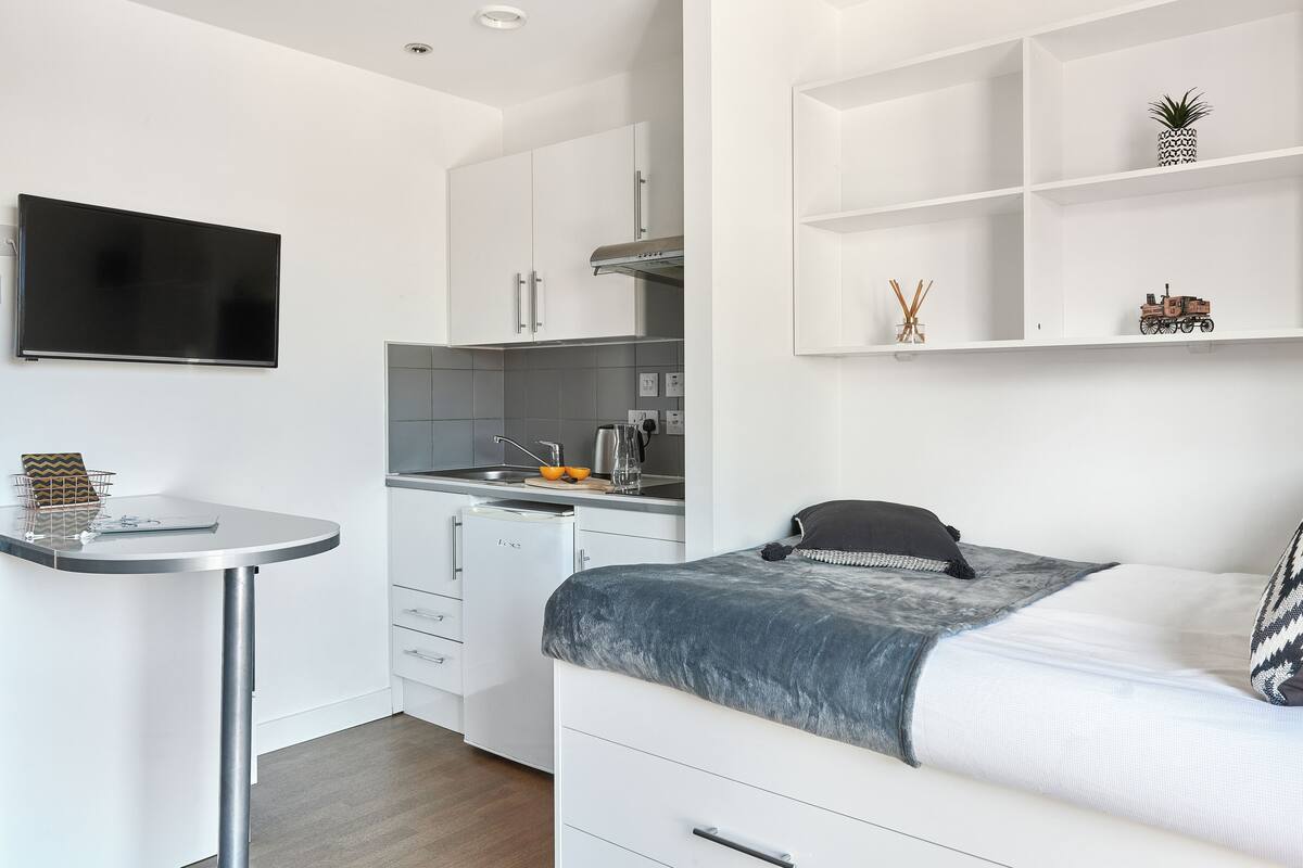 A compact, modern room with a kitchenette, a bed with a patterned pillow, a small table with a chair, and a TV mounted on the wall.