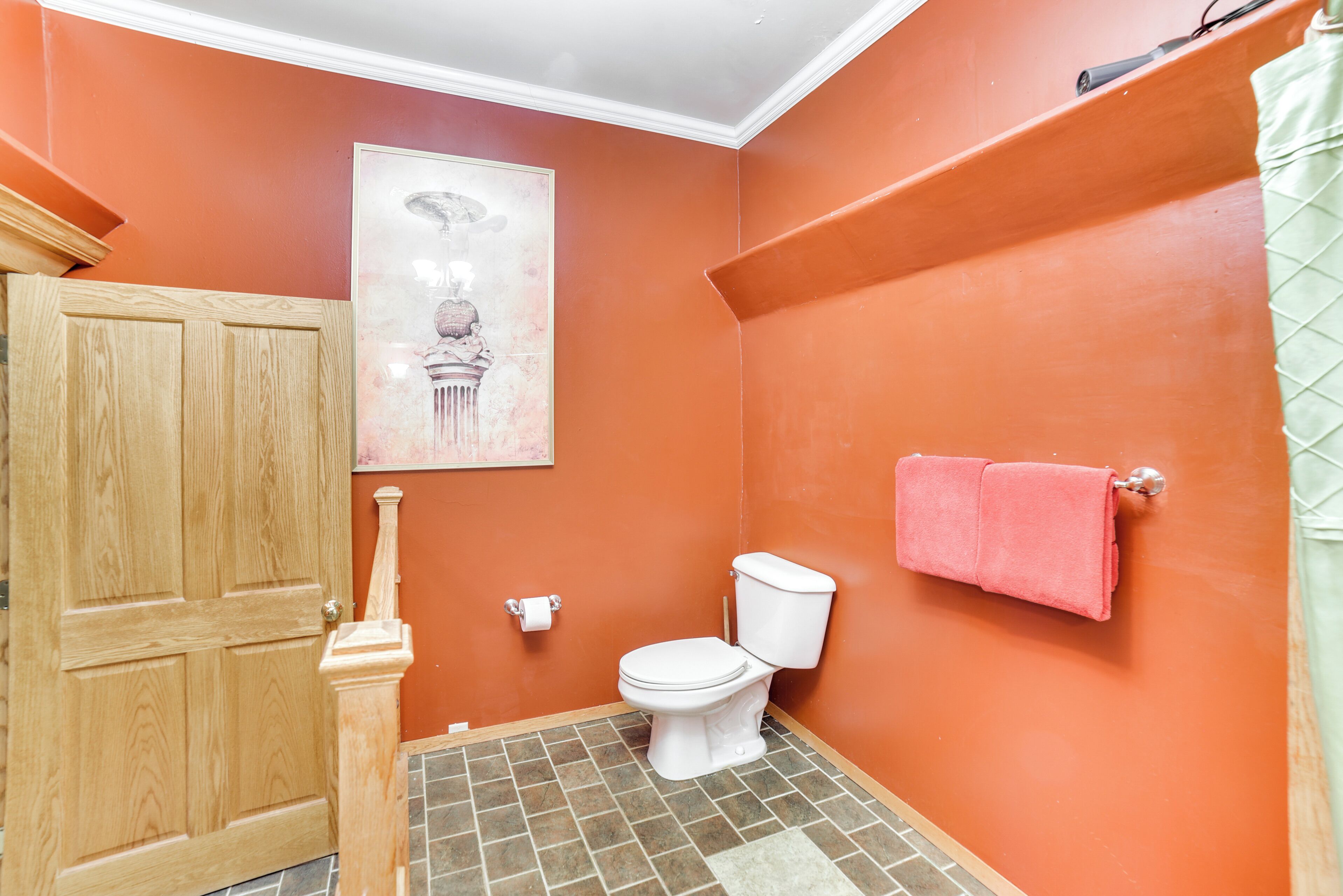 A bathroom with a wooden door, a toilet, a pink towel, and a framed picture on the wall.