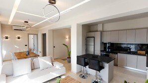 A modern kitchen with grey cabinets, a black countertop, and stainless steel appliances. There is a dining area with a white sofa and a wooden table.