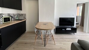 A modern kitchen with black cabinets, a microwave, and a patterned backsplash. There's a dining table with chairs in the center, a flat-screen TV on a stand, and a sofa in the living area.