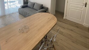 A modern living room with a wooden dining table, clear glass chairs, a grey sofa, and a ceiling fan.