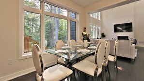 A dining area with a glass table, white chairs, and a view of trees through large windows.