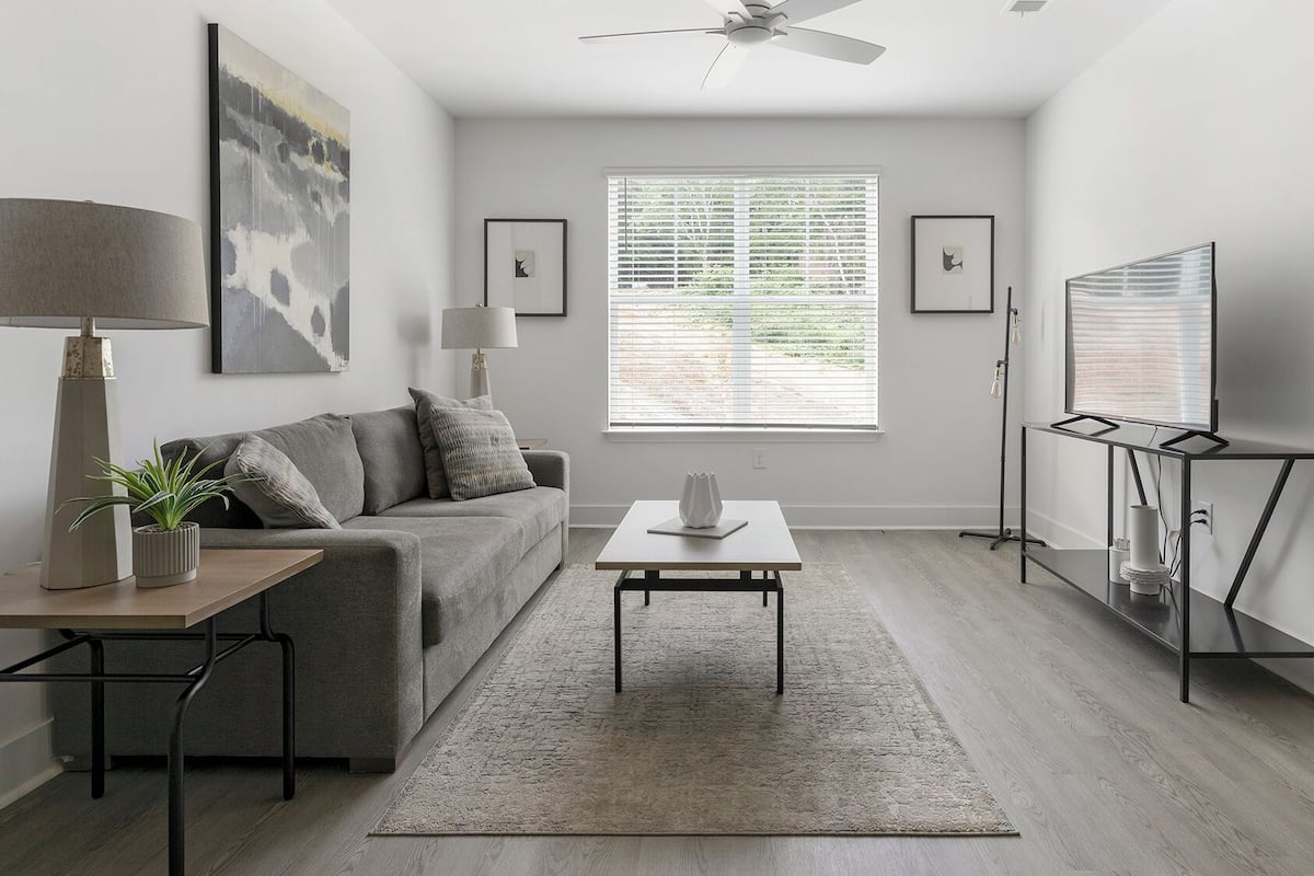 Un salon moderne avec un canapé gris, une table basse, un lampadaire, un téléviseur posé sur un meuble et une fenêtre équipée de stores.