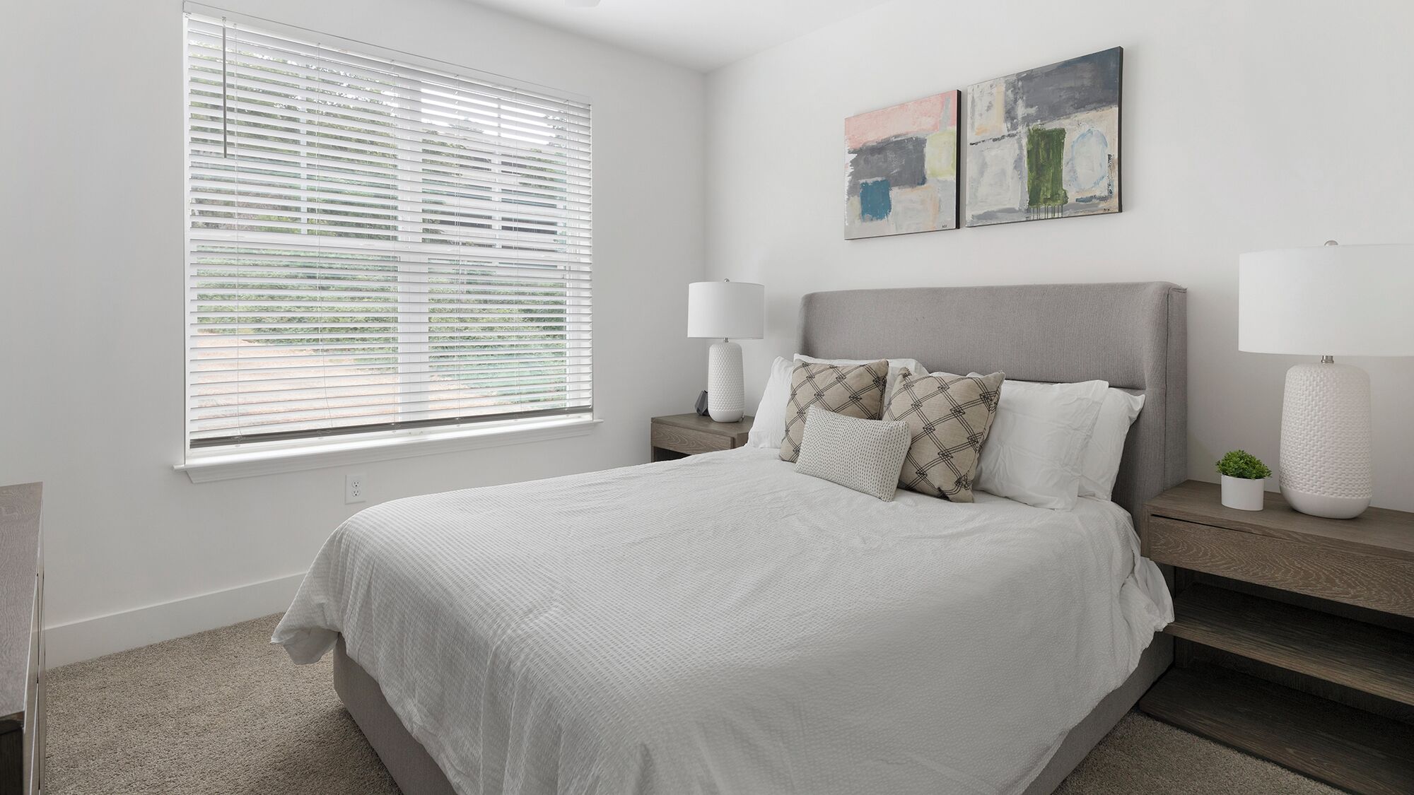 A bedroom with a large bed, a nightstand, a lamp, a window with blinds, and two abstract paintings on the wall.