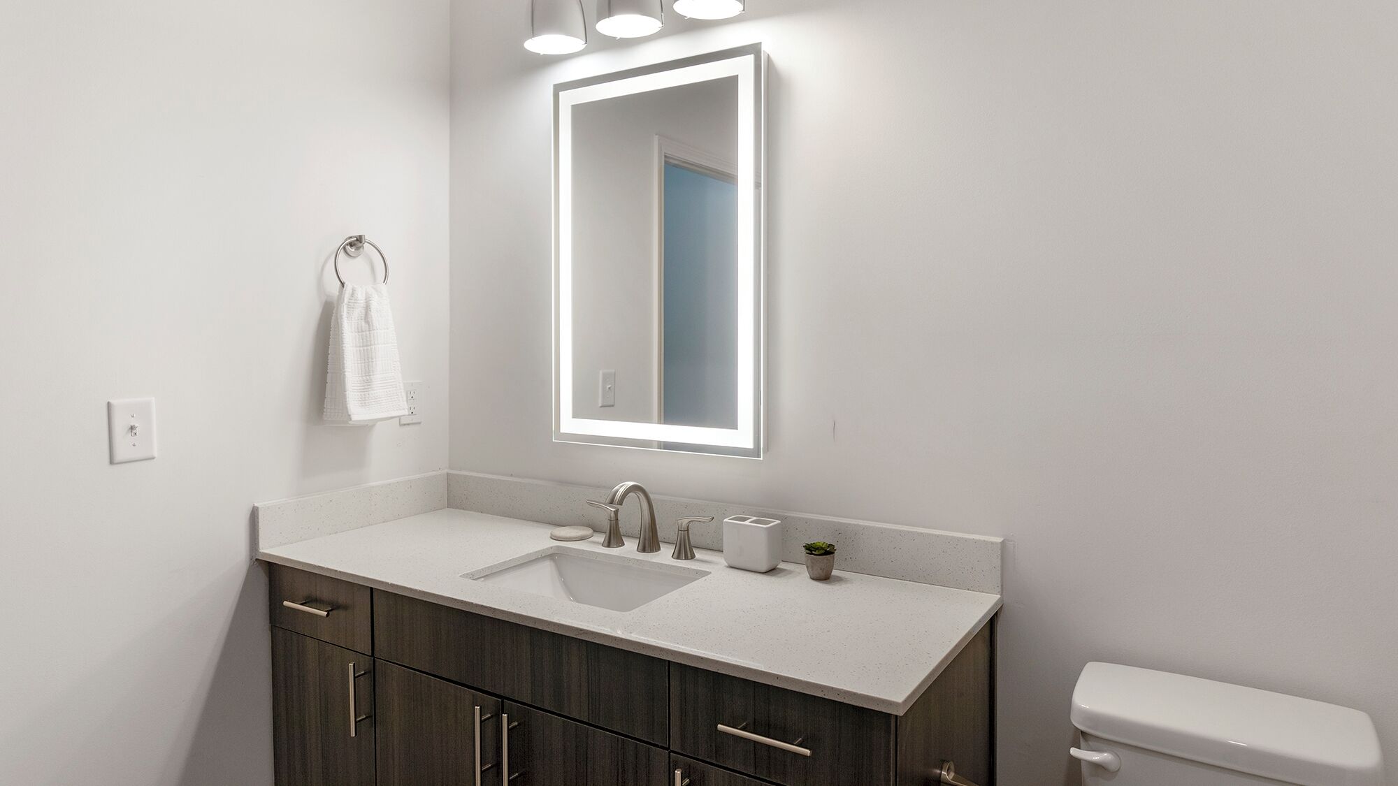 A modern bathroom with a white vanity, a large mirror with integrated lighting, a toilet, and a small plant on the counter.