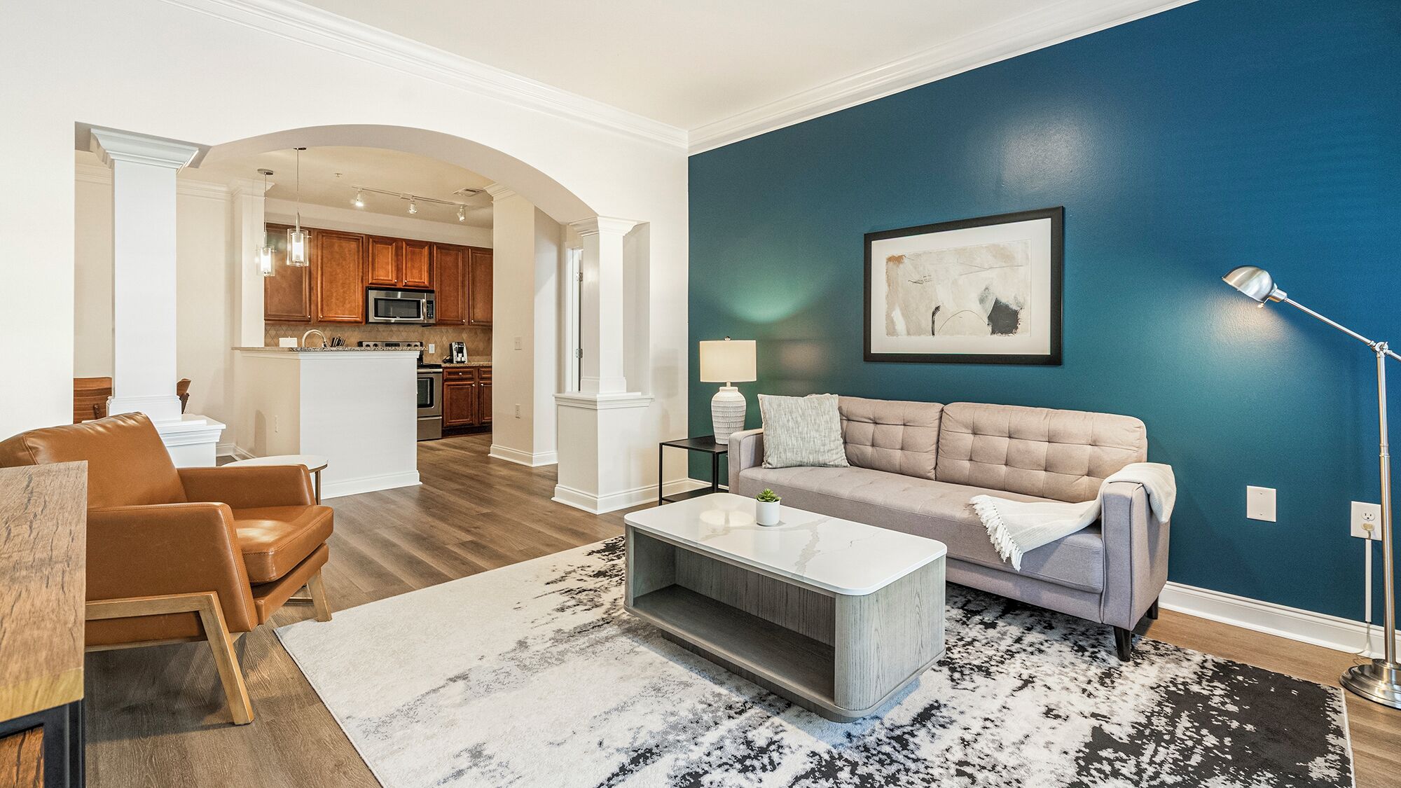 A living room with a sofa, coffee table, and a rug. There is a kitchen in the background with wooden cabinets and a microwave.