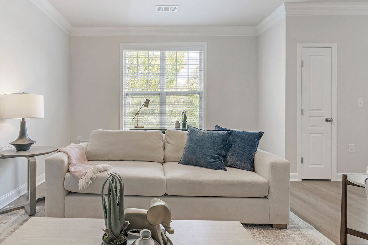 Un salón con un sofá color beige, una mesita de noche con lámpara, una planta en maceta y una ventana con persianas.
