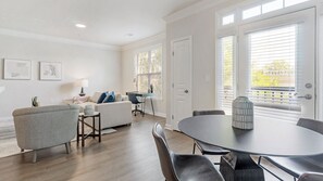 A modern living room with a grey sofa, a coffee table, and a dining area with a round table and chairs. There are two doors and windows with blinds.
