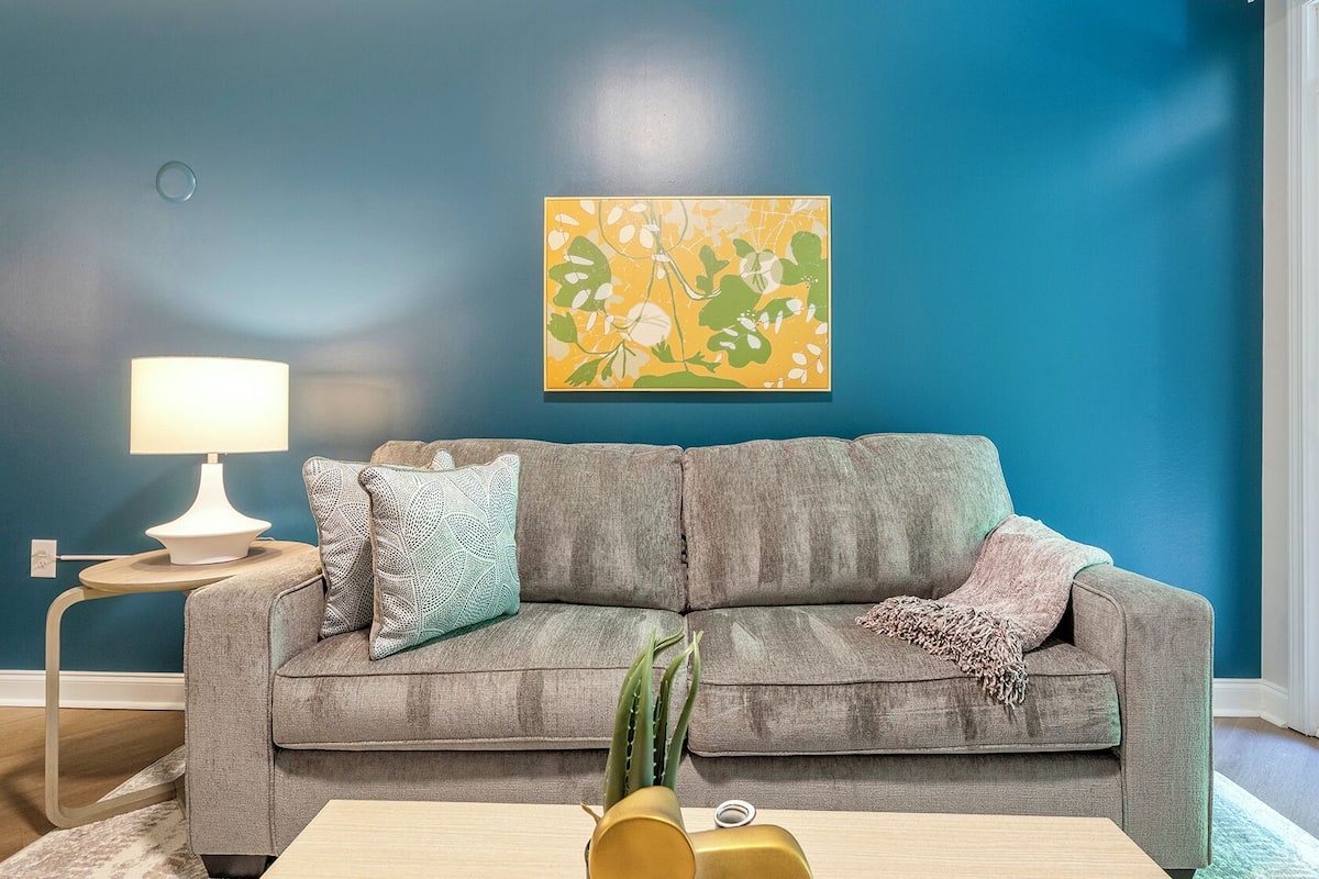 Una sala de estar con un sofá gris, una mesita de noche con lámpara y un cuadro abstracto de colores en la pared.