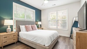 A bedroom with a bed, two bedside tables, a TV, and two windows with blinds.