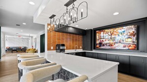 A modern kitchen with a long island, bar stools, and a large screen displaying a scenic outdoor view. - Cary Apartments by Landing (Cary)