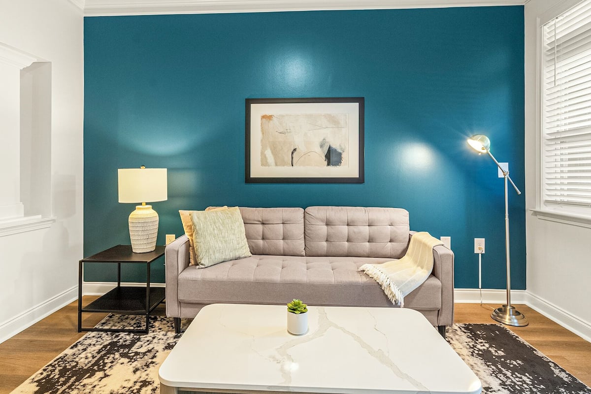 Una sala de estar con una pared verde azulado, un sofá gris, una mesa de centro blanca, una lámpara de pie y un cuadro enmarcado.