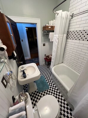Un baño con suelo de cuadros, un lavamanos blanco, un inodoro y una bañera. Hay un perchero con toallas y una cesta en la pared.