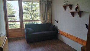 Una habitación con suelo de madera, sofá verde oscuro, un armario de madera y una ventana con vistas a árboles y montañas.