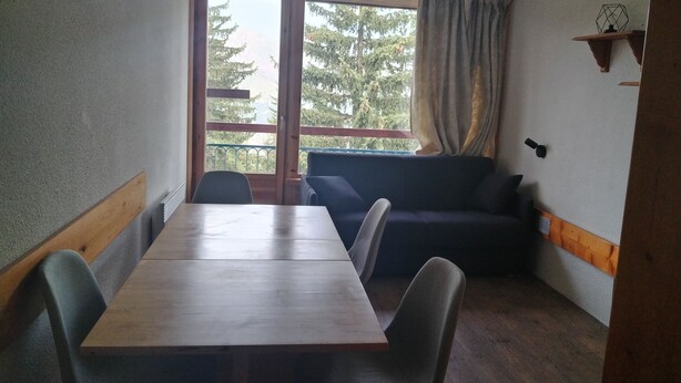 Una habitación con una mesa y sillas de madera, un sofá oscuro, una ventana con cortinas y un estante con un objeto geométrico.