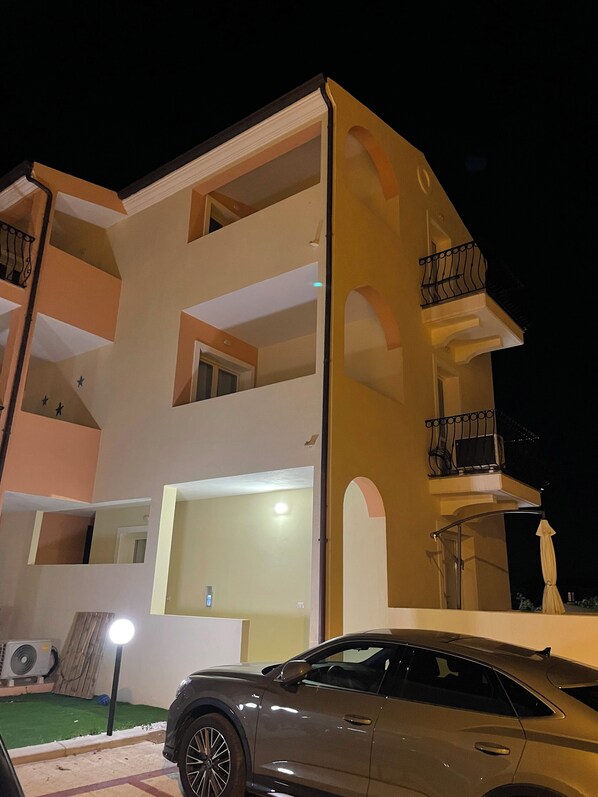 Un edificio de varios pisos con balcones, un automóvil estacionado adelante y una pequeña área ajardinada.