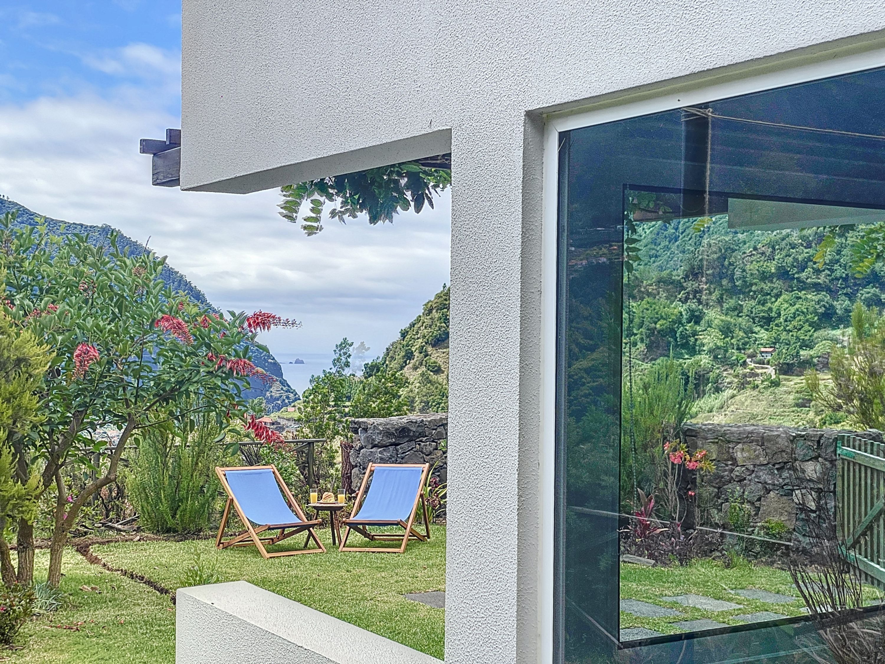 A modern outdoor area with two lounge chairs and a table, overlooking a lush garden and a mountainous landscape.