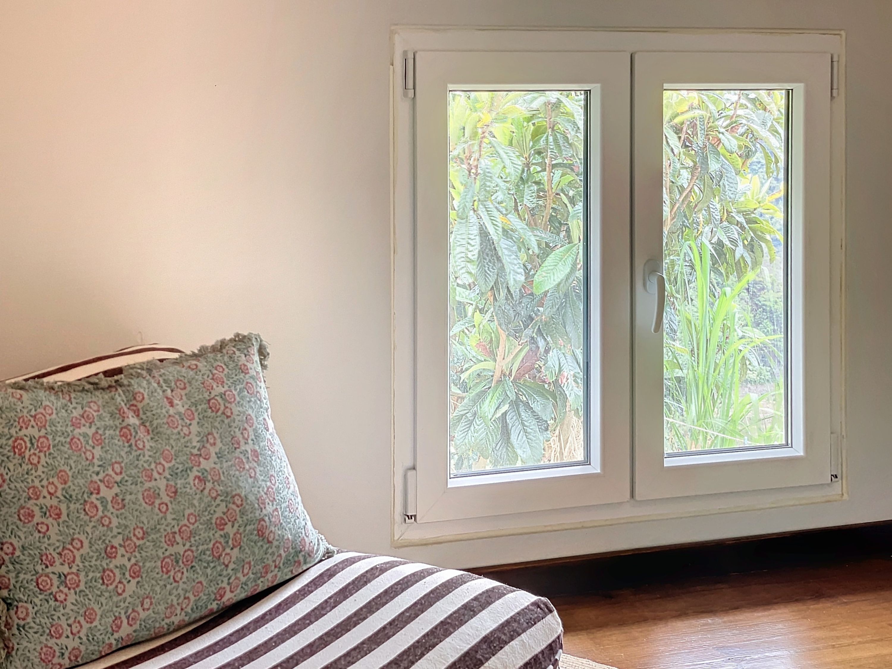 A room with a bed, a floral pillow, and a window with a view of greenery.