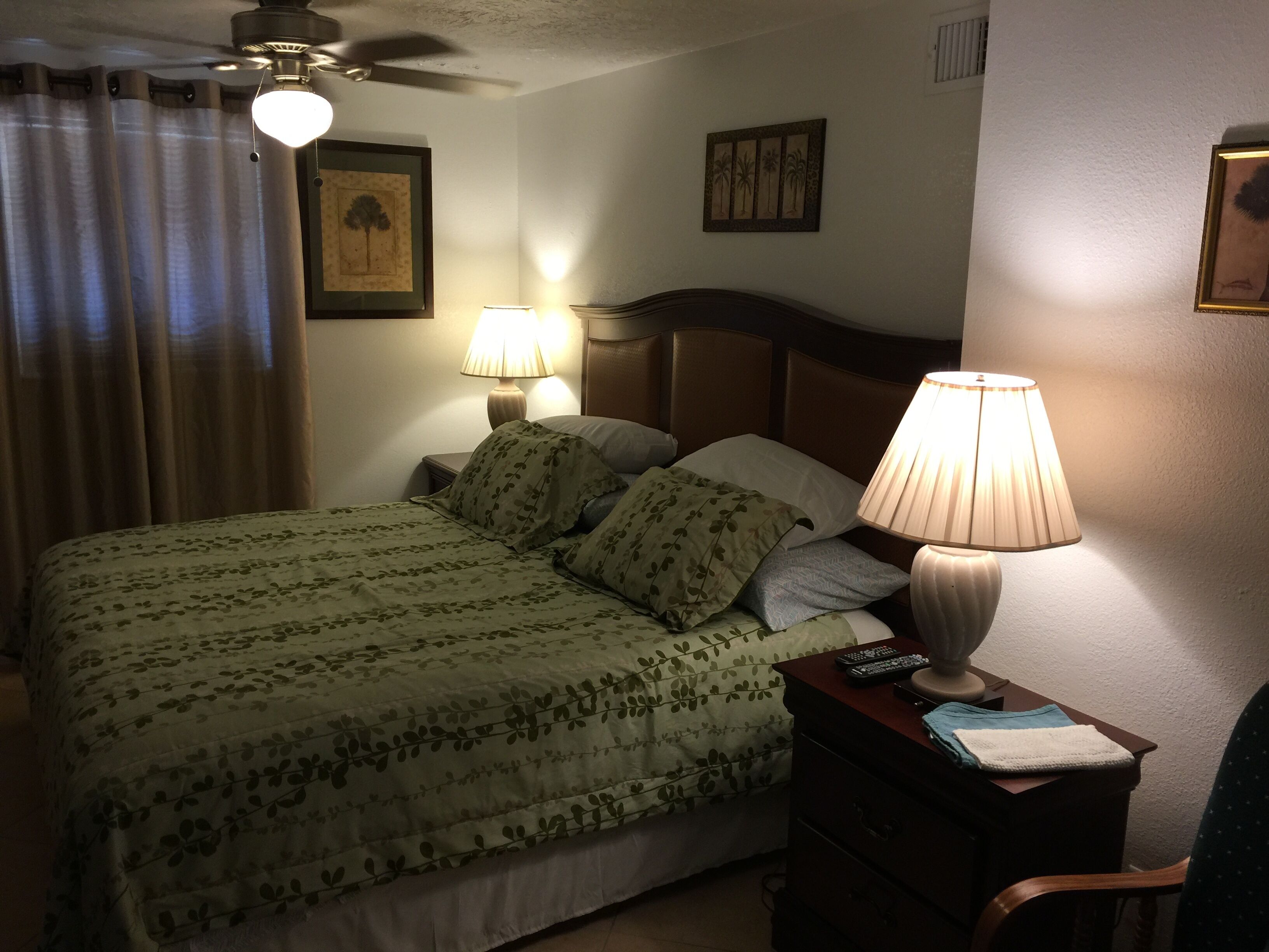 A neatly made bed with floral bedding, two bedside lamps, a ceiling fan, and a small table with a remote control.