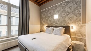 Habitación de hotel con una cama grande, una mesita de noche, una lámpara de pared y una ventana con cortinas.