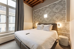 Quarto de hotel com uma cama grande, mesa de cabeceira, luminária na parede e janela com cortinas.