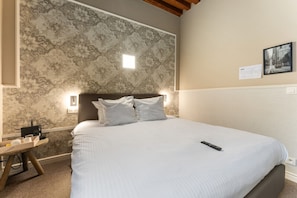 Uma cama bem arrumada com lençóis brancos, uma mesa de cabeceira com abajur, e uma parede com papel de parede estampado e um quadro.