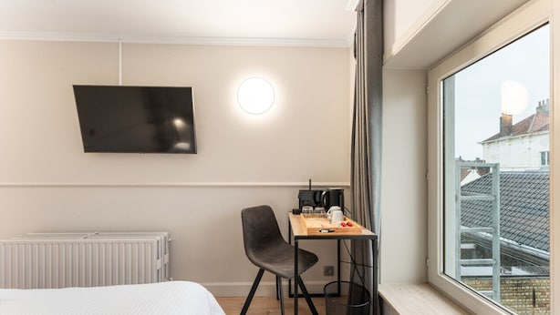 Une chambre d’hôtel comprenant un lit, un bureau avec une chaise, un téléviseur fixé au mur et une fenêtre donnant sur des immeubles.